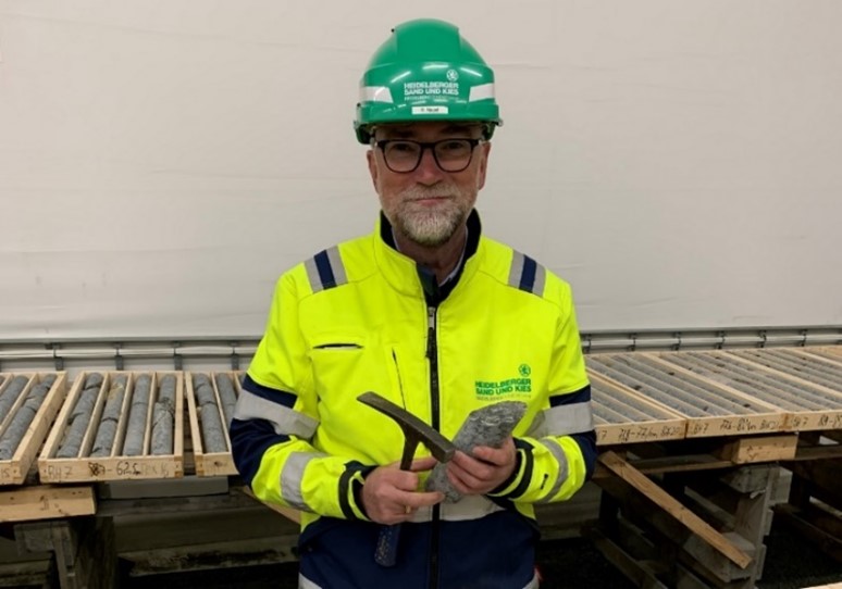Man with yellow protective jacket and green helmet holds a hammer and a drill core in his hands