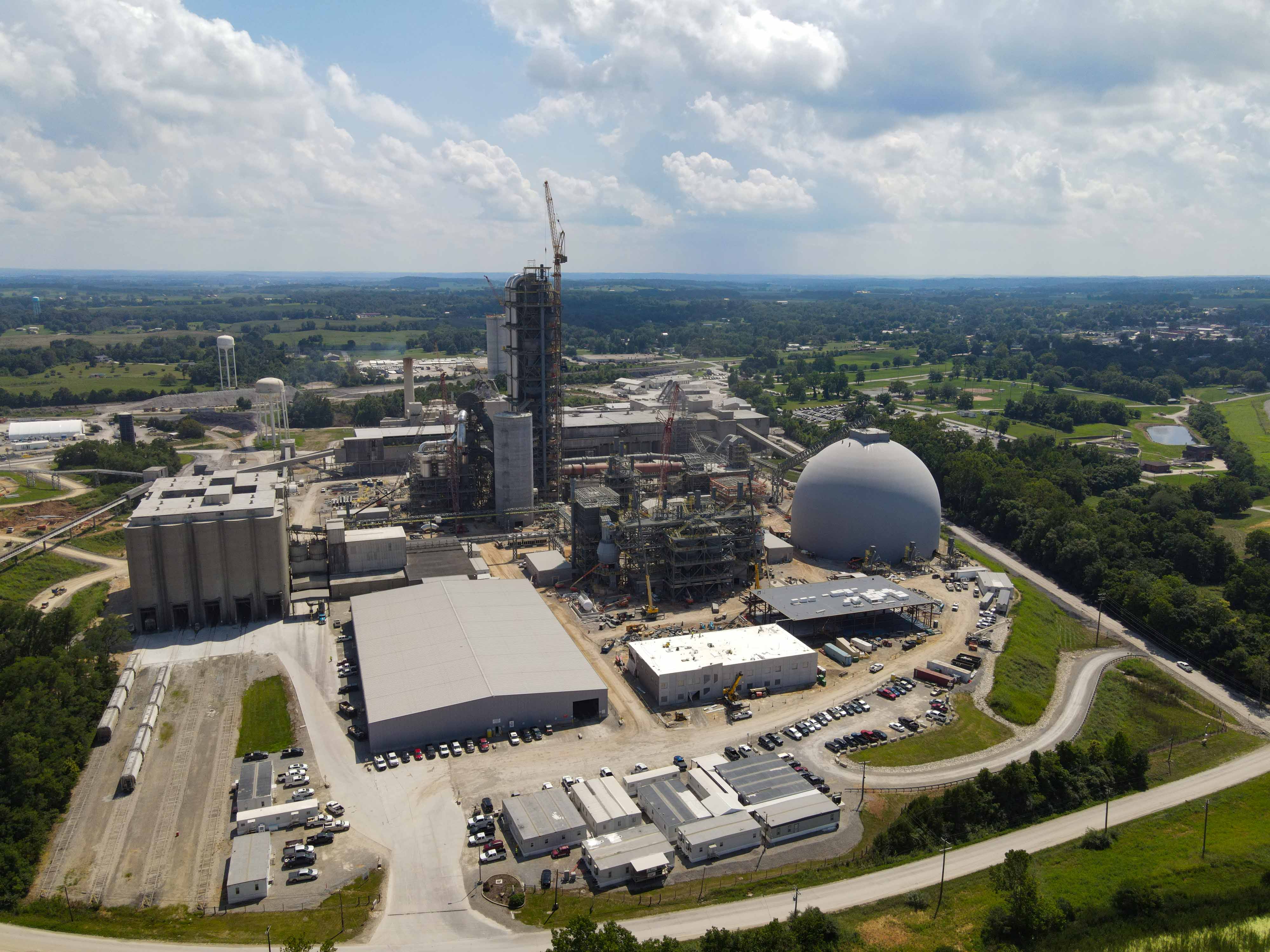Ein großer Zementwerk im Bau, mit mehreren Gebäuden und einem Silo sichtbar. Kräne und andere Baumaschinen sind vor Ort.