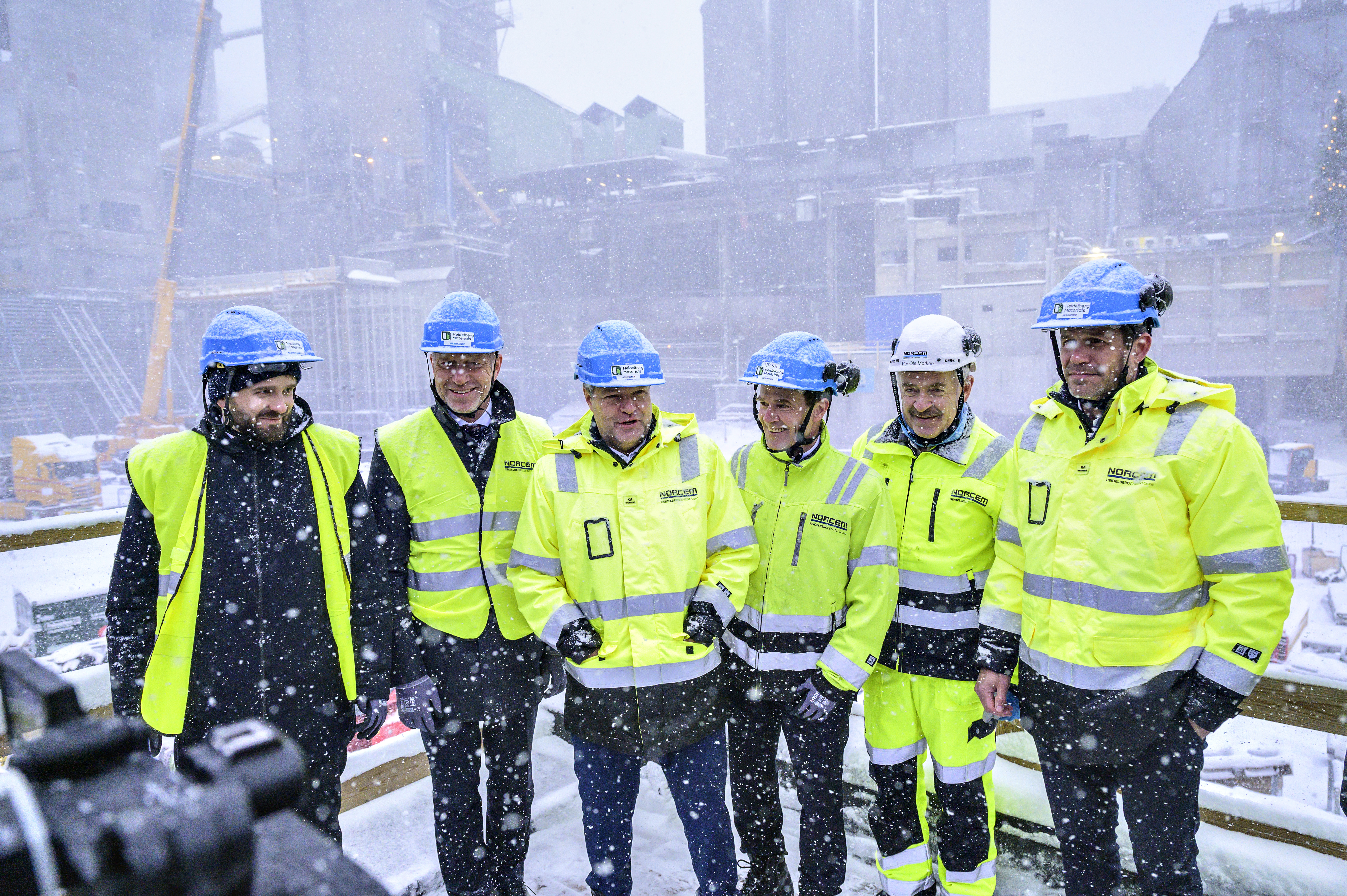 Eine Gruppe von Männern in Arbeitskleidung und mit Helmen stehen vor einem Zementwerk
