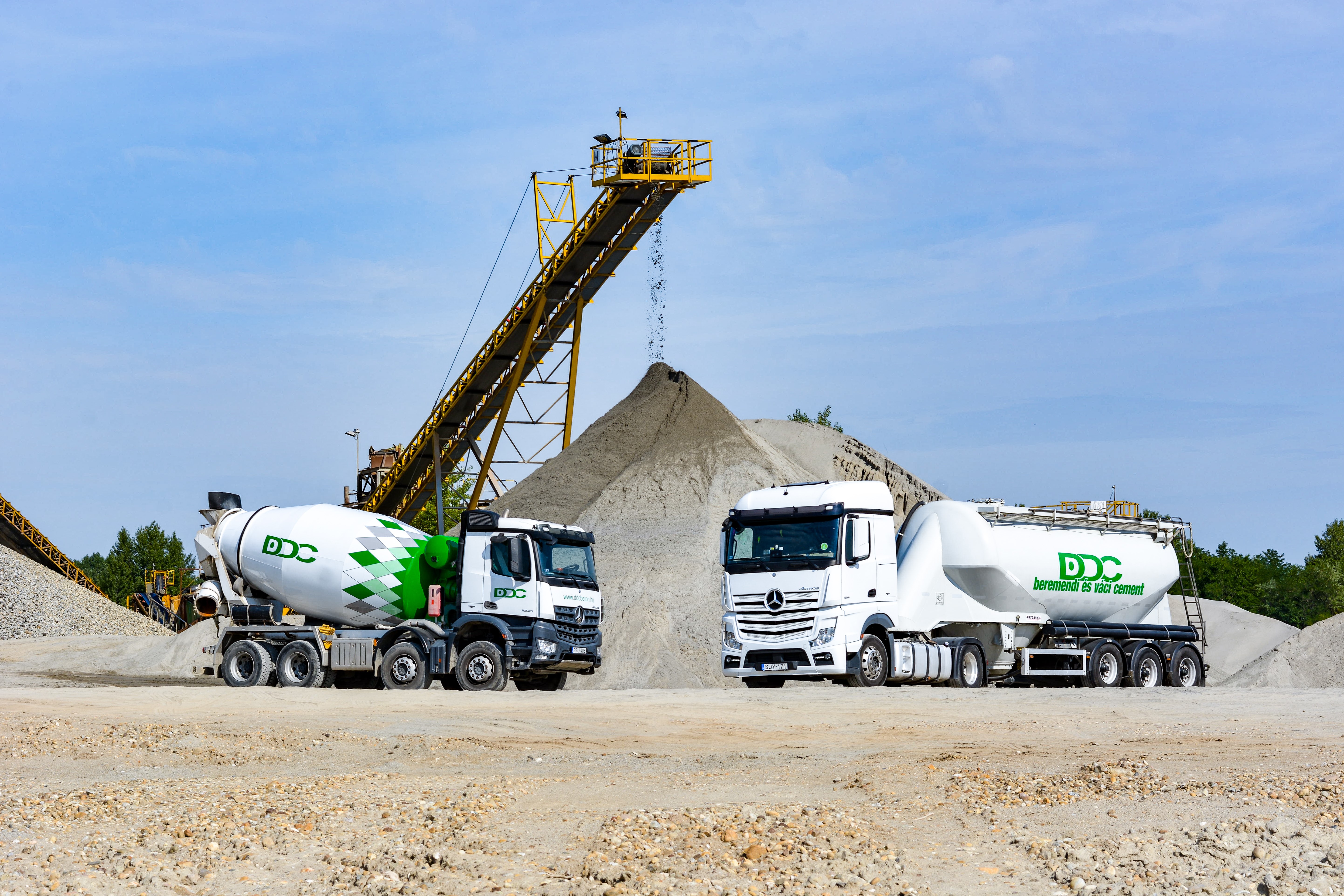 Ready-mixed truck and silo truck standing in front of a sand heap, sand trickles down from a conveyor belt