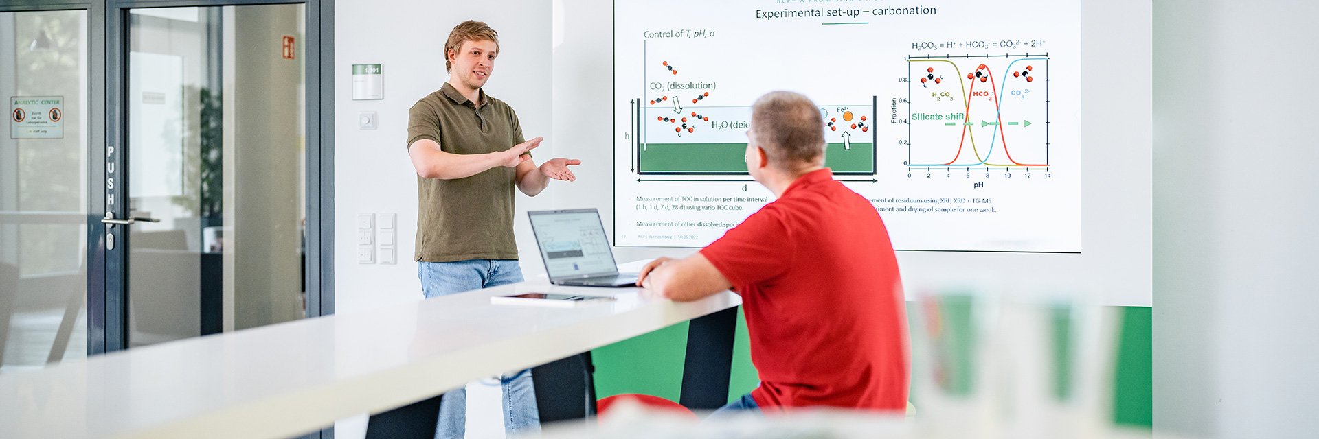 A man standing in front of a white board, explaining something to another person that is sitting on a table