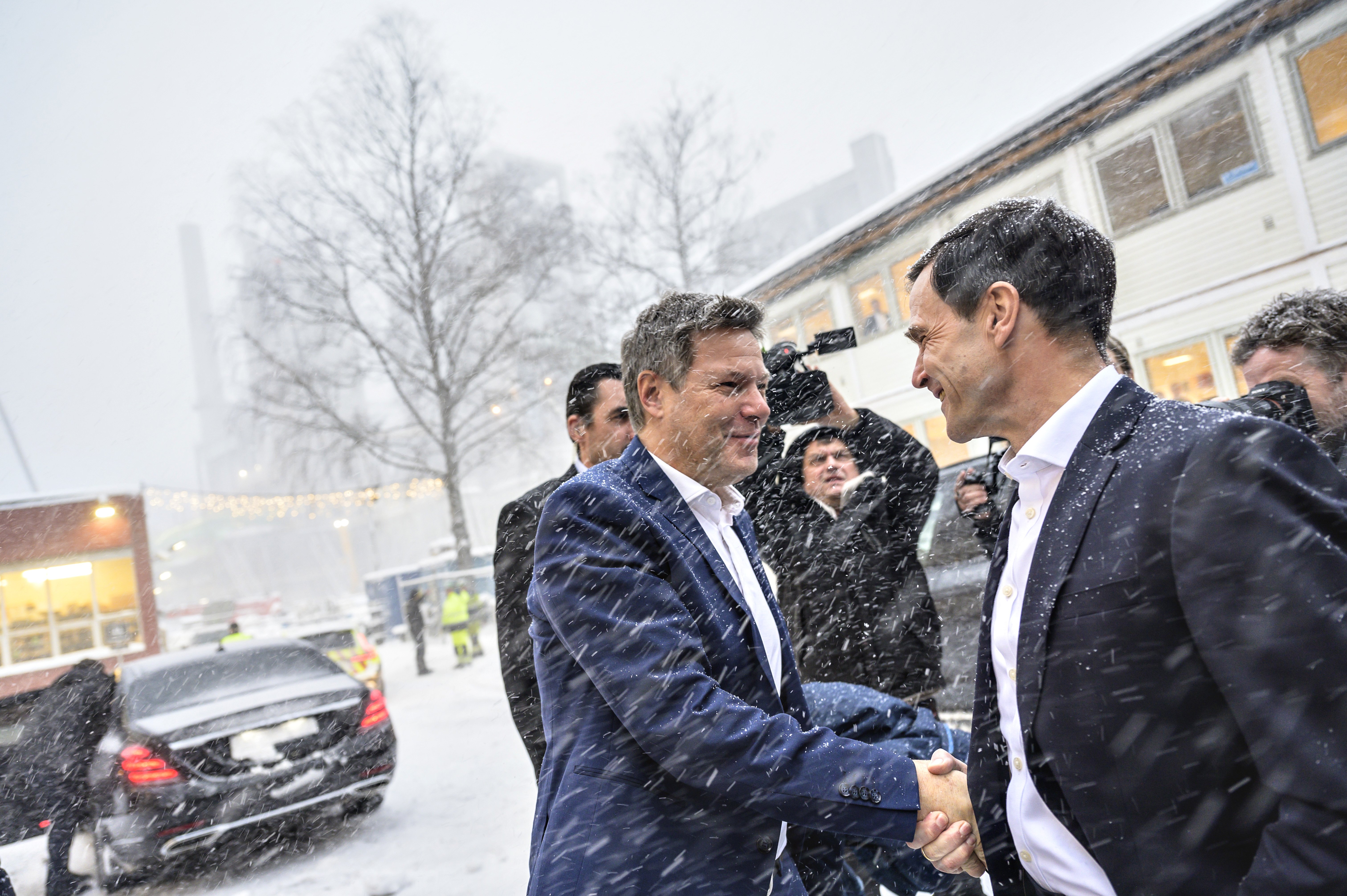 Two men in suits greet each other in the snow flurry, press people in the background