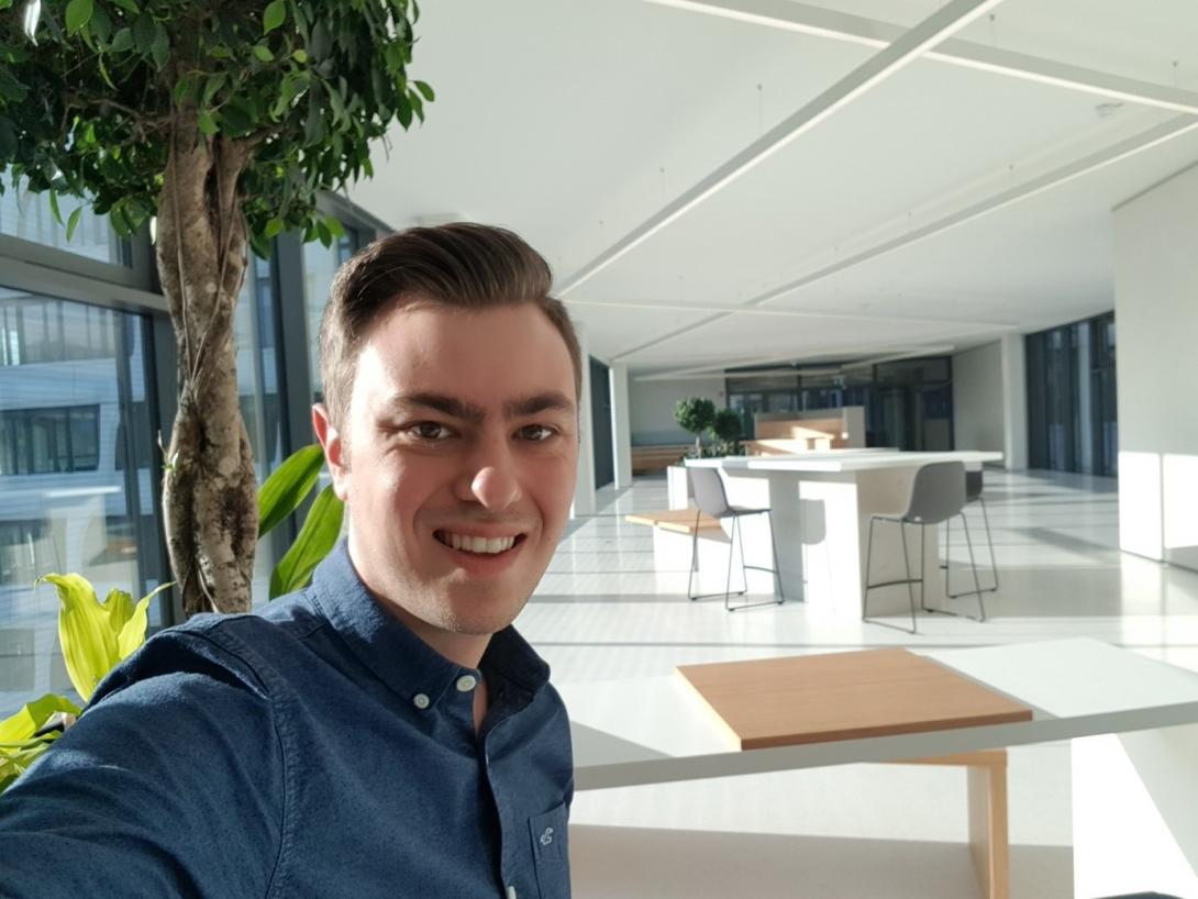 Young man in shirt in front of an area of an office building furnished with bar table and plants