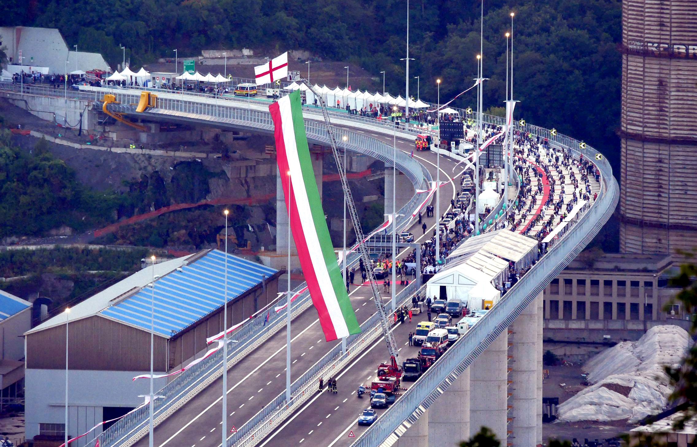 A curved bridge with a big crowd on it, the italian flag is raised by a crane which is attached to a fire truck