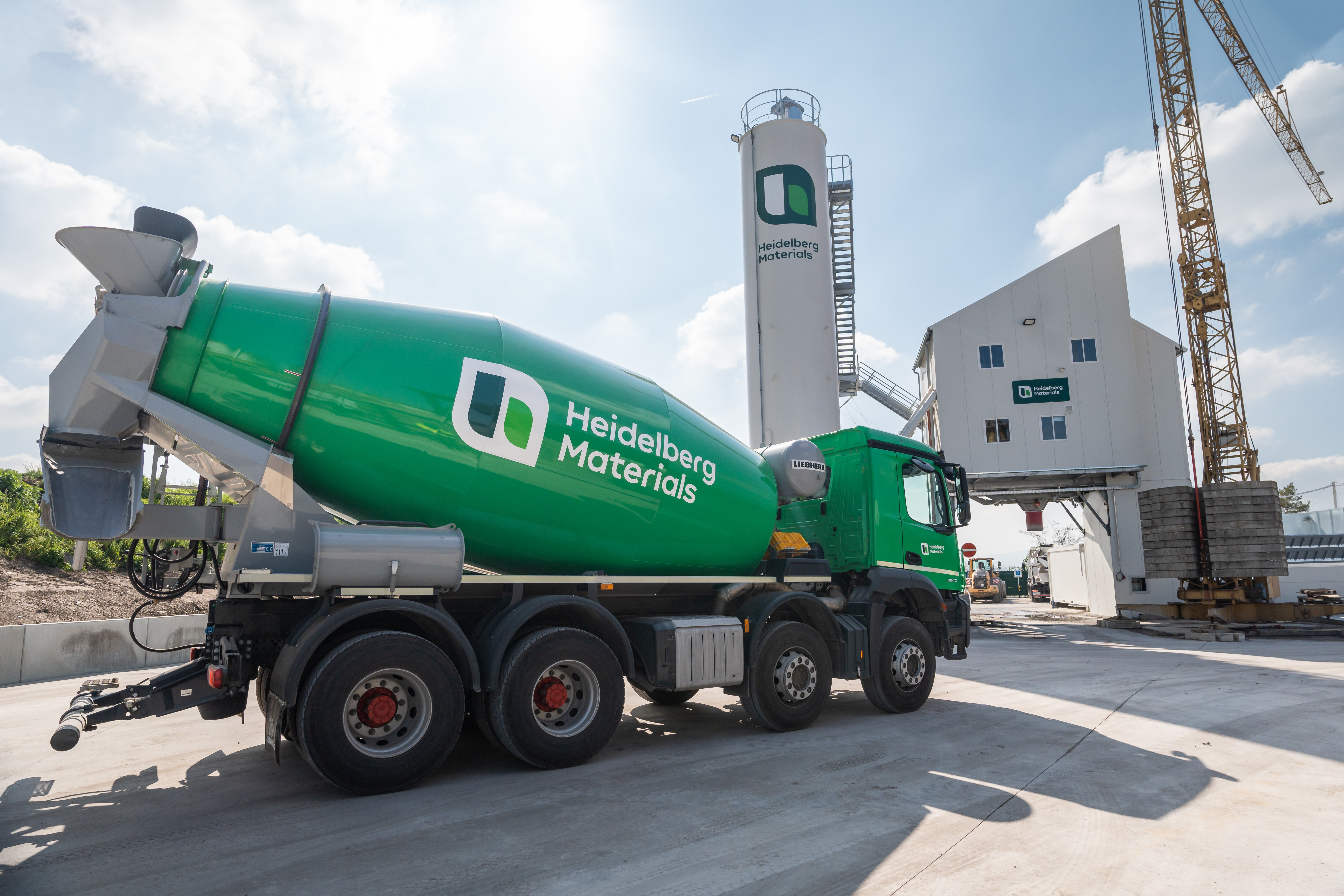Green ready-mix truck in front of a silo, an industrial building and a crane