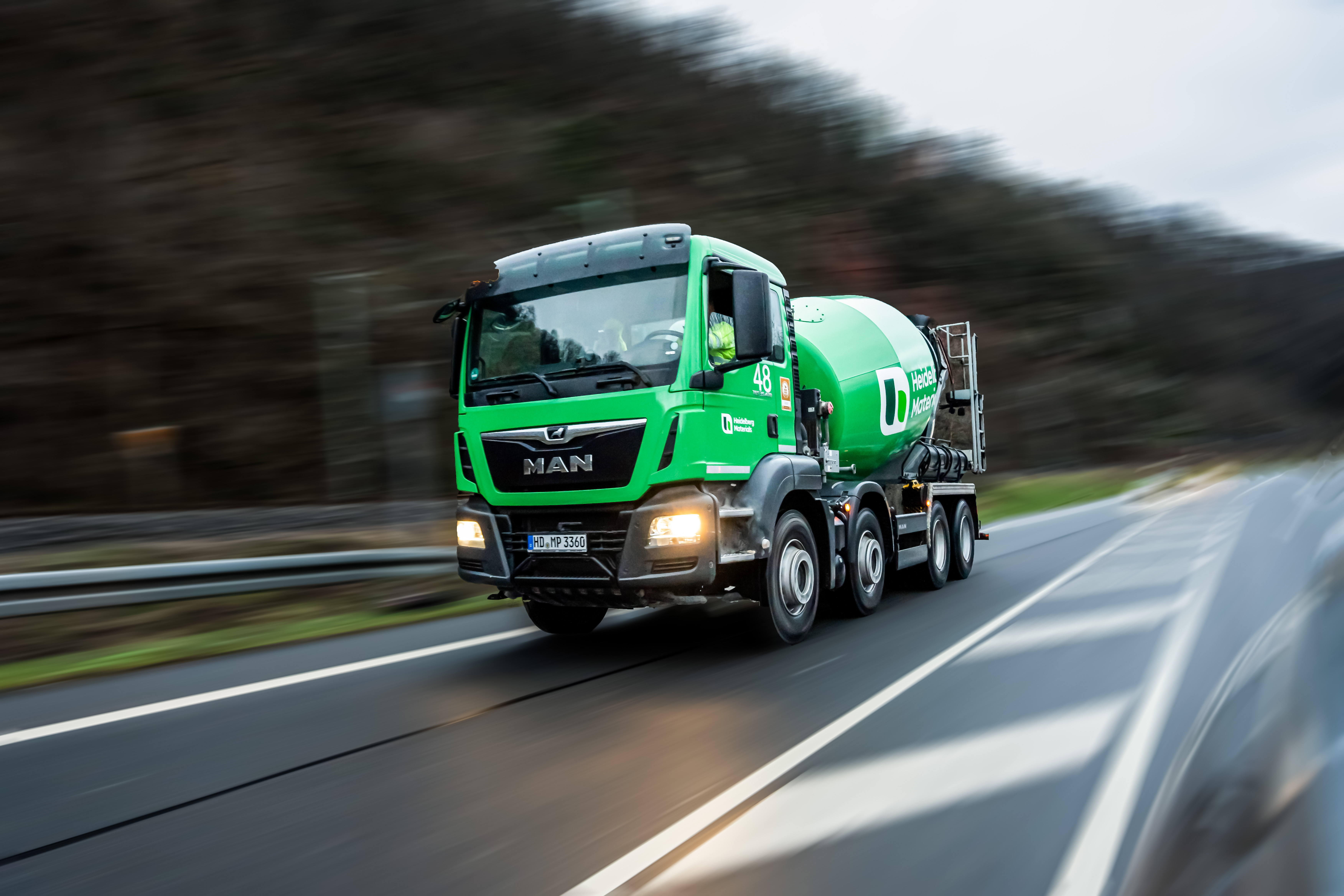 Green ready-mix truck on the road, motion blurred trees in the background