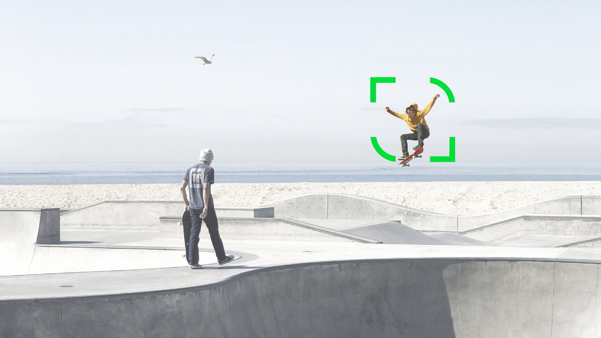 Zwei Skater in einem Skatepark aus Beton, einer springt, der andere hält sein Board. Im Hintergrund sind das Meer und eine Möwe zu sehen.