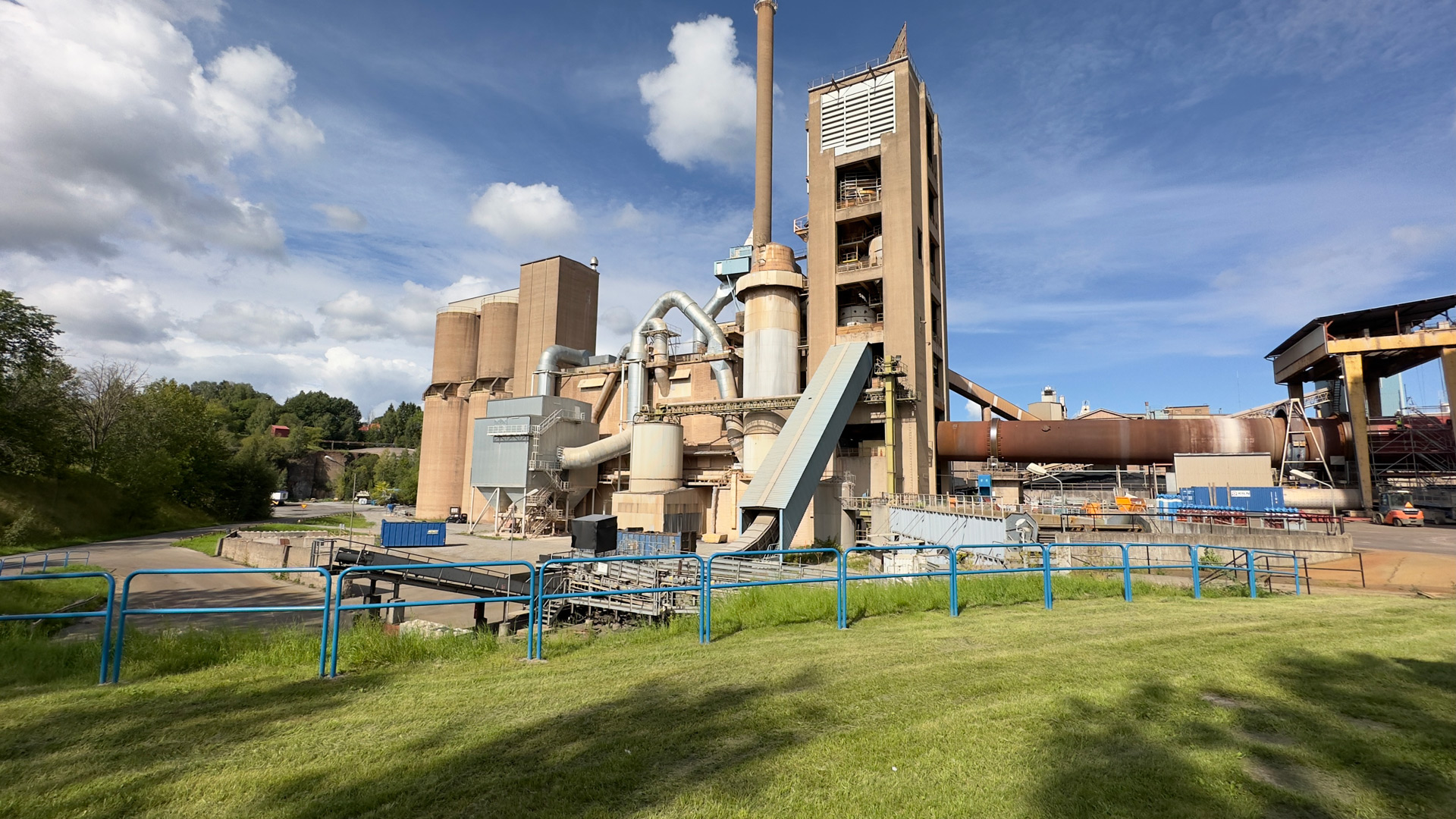 Cement plant in front of a blue sky with white clouds, in front of it a green area