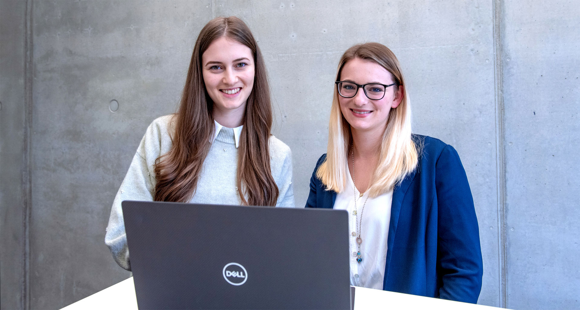 Zwei junge Frauen stehen vor einer Sichtbetonwand und blicken auf einen aufgeklappten Laptop