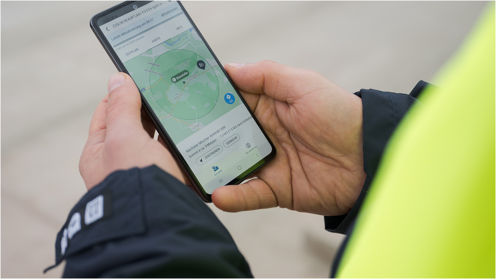Eine Person hält ein Smartphone mit einer geöffneten App, die eine Karte und einen markierten Standort anzeigt. Die Hände tragen eine dunkle Jacke, und im rechten Teil des Bildes ist eine gelbe Sicherheitsweste sichtbar. Die Karte im Smartphone zeigt einen grünen Kreis, der den Standort hervorhebt.