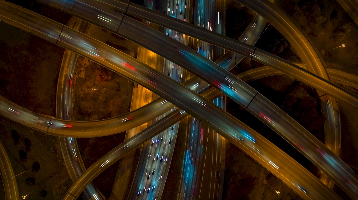 Aerial view of a complex multi-level highway interchange at night, illuminated by streetlights. Traffic flows along various curved and intersecting roadways, with red and white car lights visible in opposite directions.