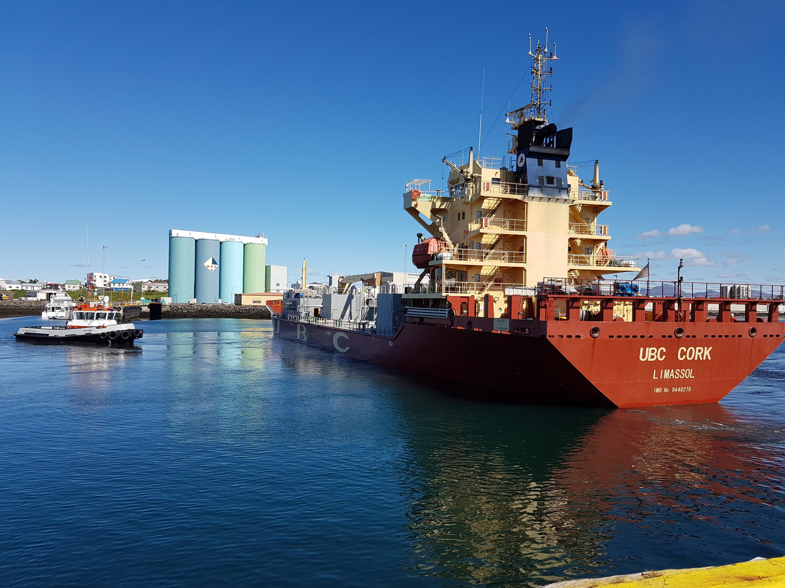 Ein rotes Frachtschiff fährt im Hafen ein, begleitet von einem kleinen Schlepperboot auf der linken Seite. Im Hintergrund sind ein Lagergebäude und der blaue Himmel zu sehen. Im Vordergrund eine gelb gestrichene Kaimauer.