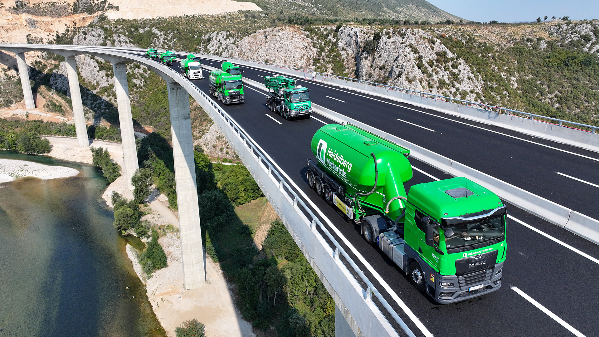 Grüne Betonmischer-Lkws fahren auf einer modernen Brücke, die über eine Schlucht mit Fluss führt. Im Hintergrund sind Berge und Natur sichtbar. Die Fahrzeuge sind in einer Kolonne angeordnet und zeigen ein firmenspezifisches Design.