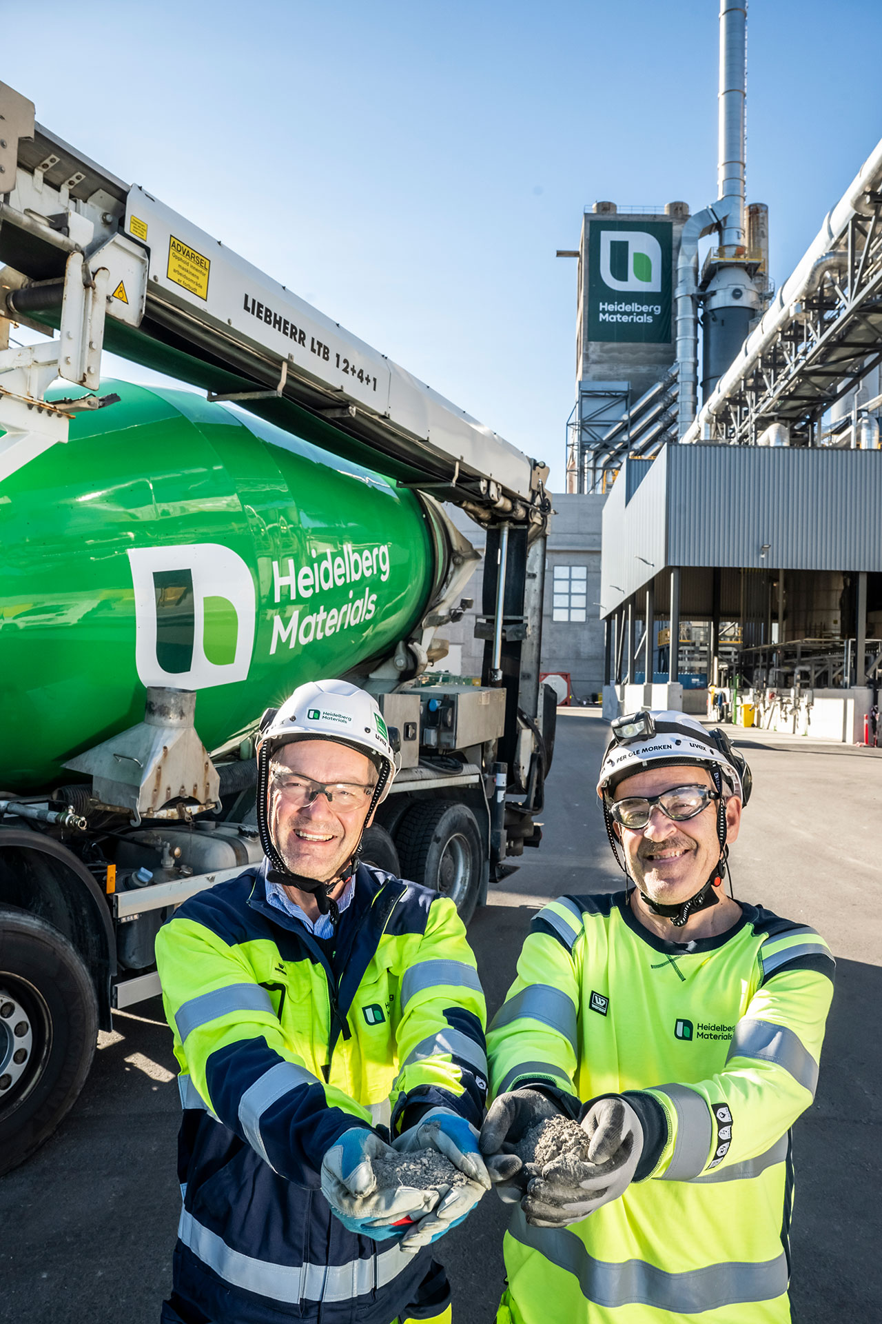 Two Heidelberg Materials workers holding cement in front of a ready-mixed concrete truck and a cement plant.