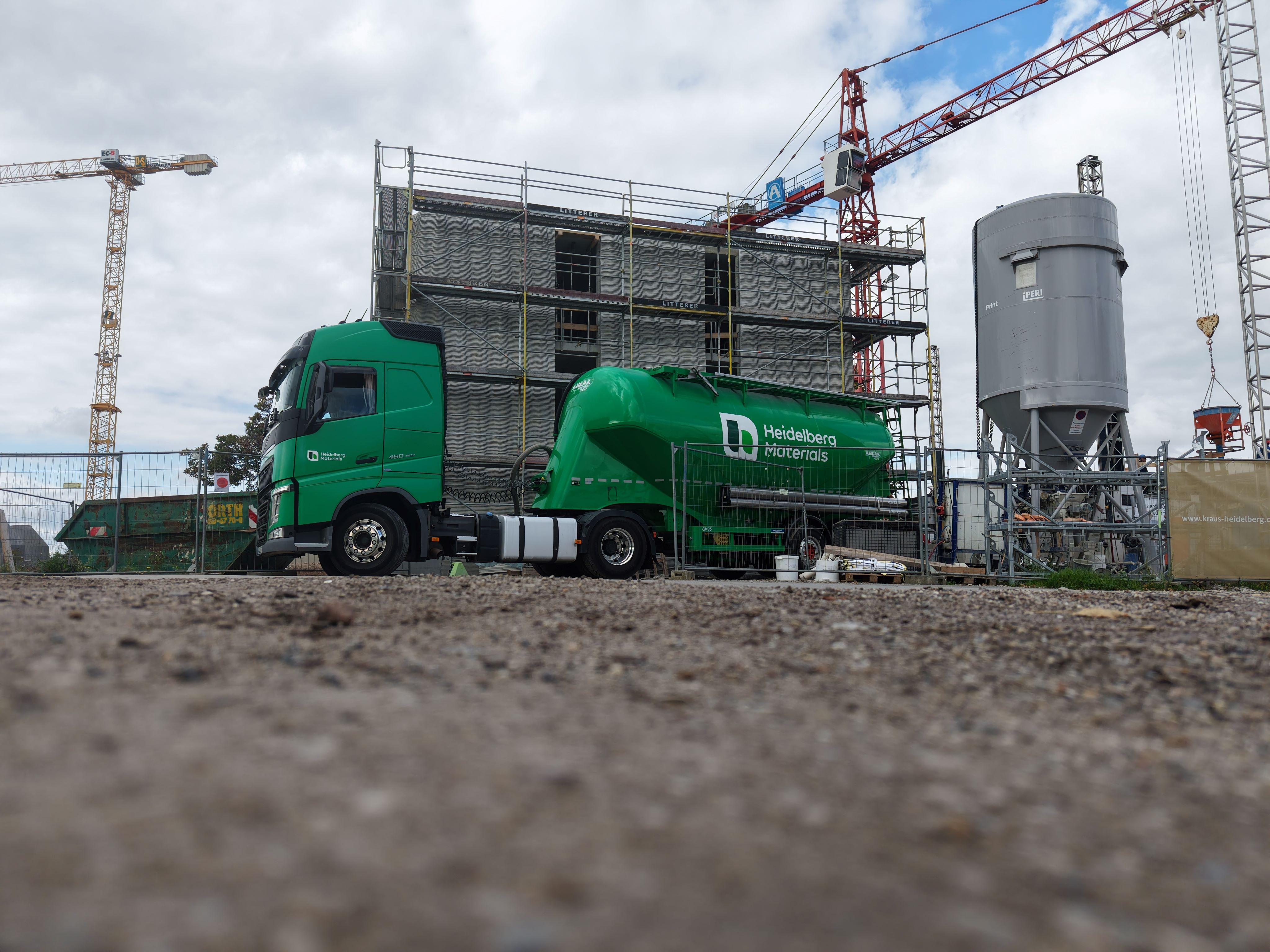 Grüner Lastwagen mit Anhänger auf einer Baustelle, im Hintergrund ein mehrstöckiger Rohbau mit Gerüsten, zwei Baustellenkräne und ein großer Silobehälter, wolkiger Himmel darüber.