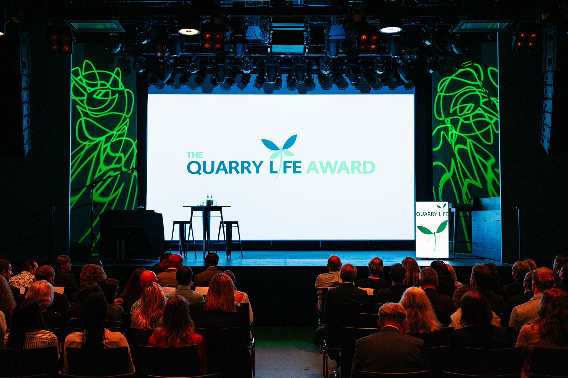 Bühne mit großer Leinwand, auf der "The Quarry Life Award" steht; davor zwei leere schwarze Rednerpulte und ein Rednerpult rechts mit Logo; Publikum sitzt in mehreren Reihen, Hintergrund mit grünen, beleuchteten Linienmustern.