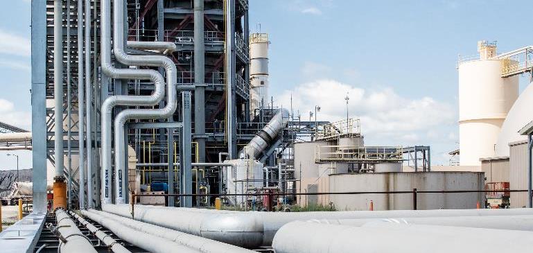 Industrieanlage mit großen Rohrleitungen im Vordergrund, verbunden mit einem hohen metallischen Gebäudestruktur links. Im Hintergrund sind runde Tanks und weitere Industriebauteile sichtbar, bei klarem Himmel und Tageslicht.