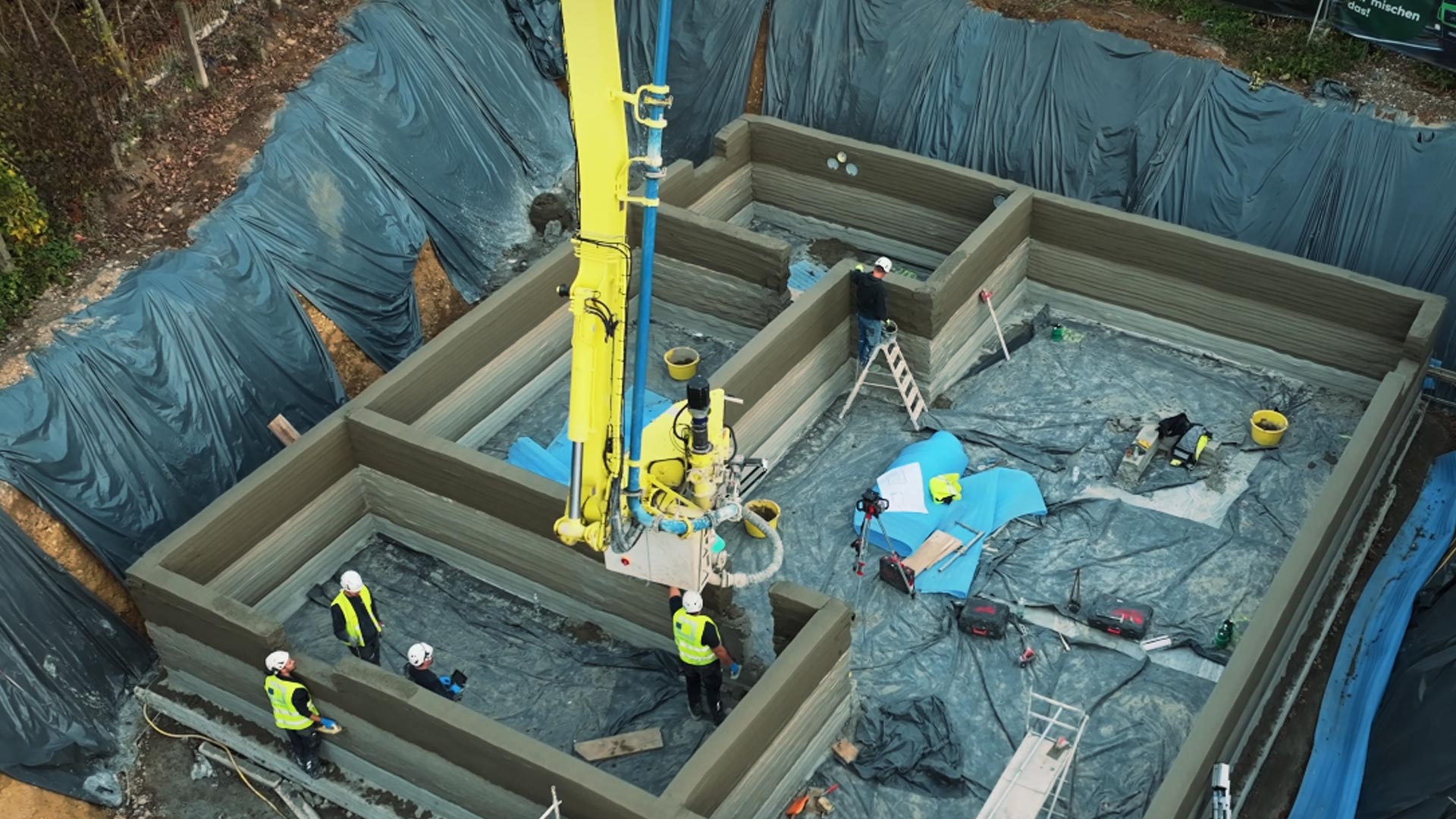 Baustelle, auf der Arbeiter eine 3D-gedruckte Betonkonstruktion erstellen. An einem gelben Kran in der Mitte hängen Geräte.