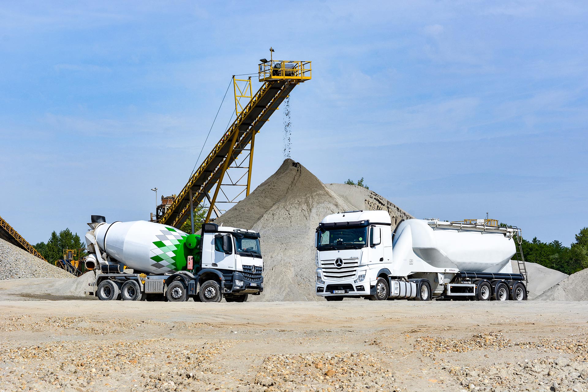Ready-mixed truck and silo truck standing in front of a sand heap, sand trickles down from a conveyor belt