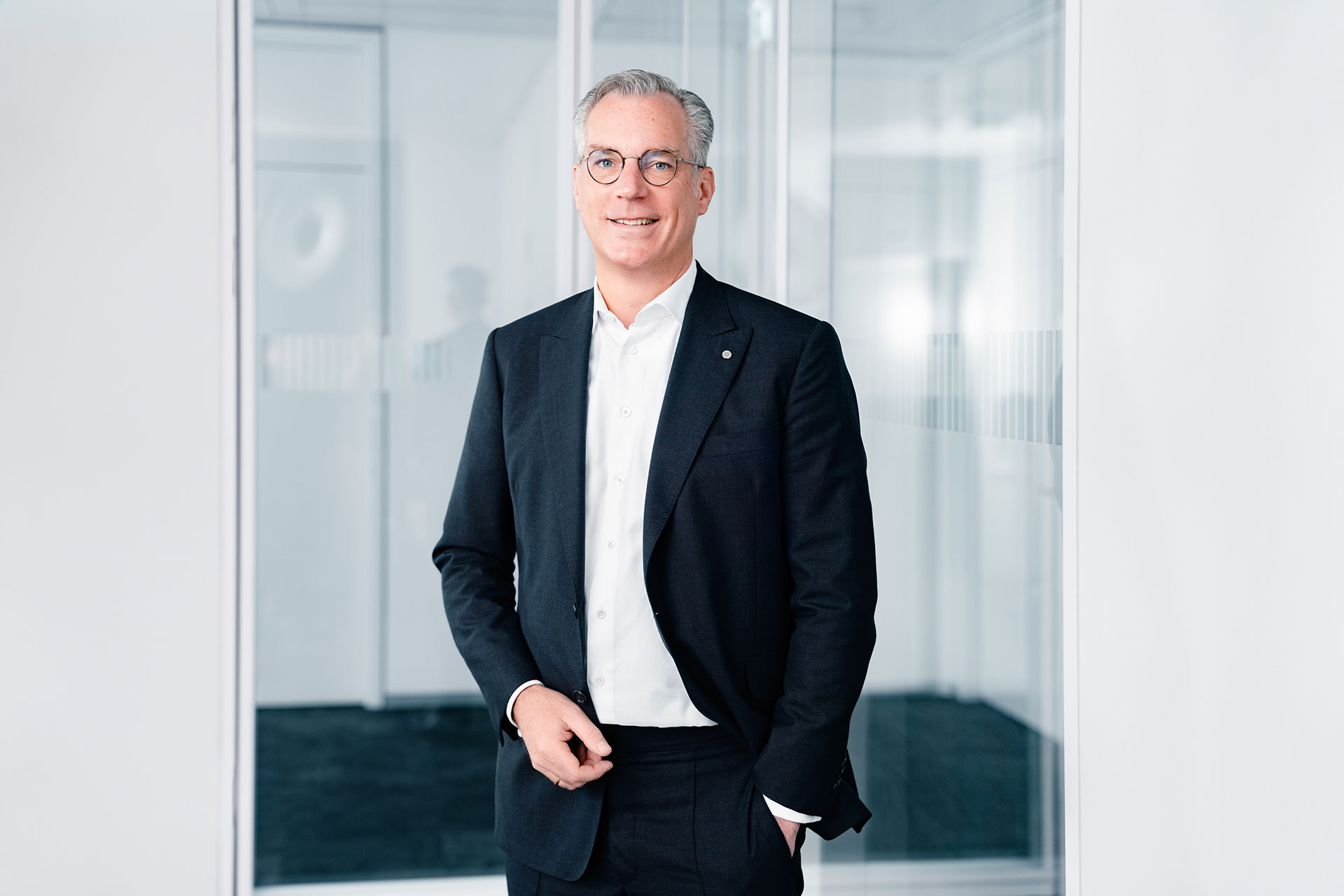 Gunnar Groebler wearing a black suit and white shirt standing in a modern office environment with glass walls.