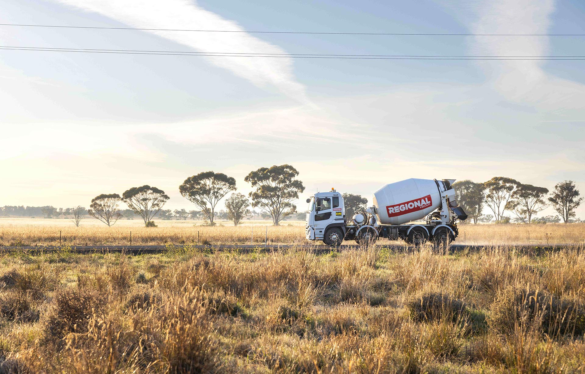A ready-mixed concrete truck drives through a rural landscape with tall dry grass in the foreground and scattered trees under a lightly clouded sky in the background.