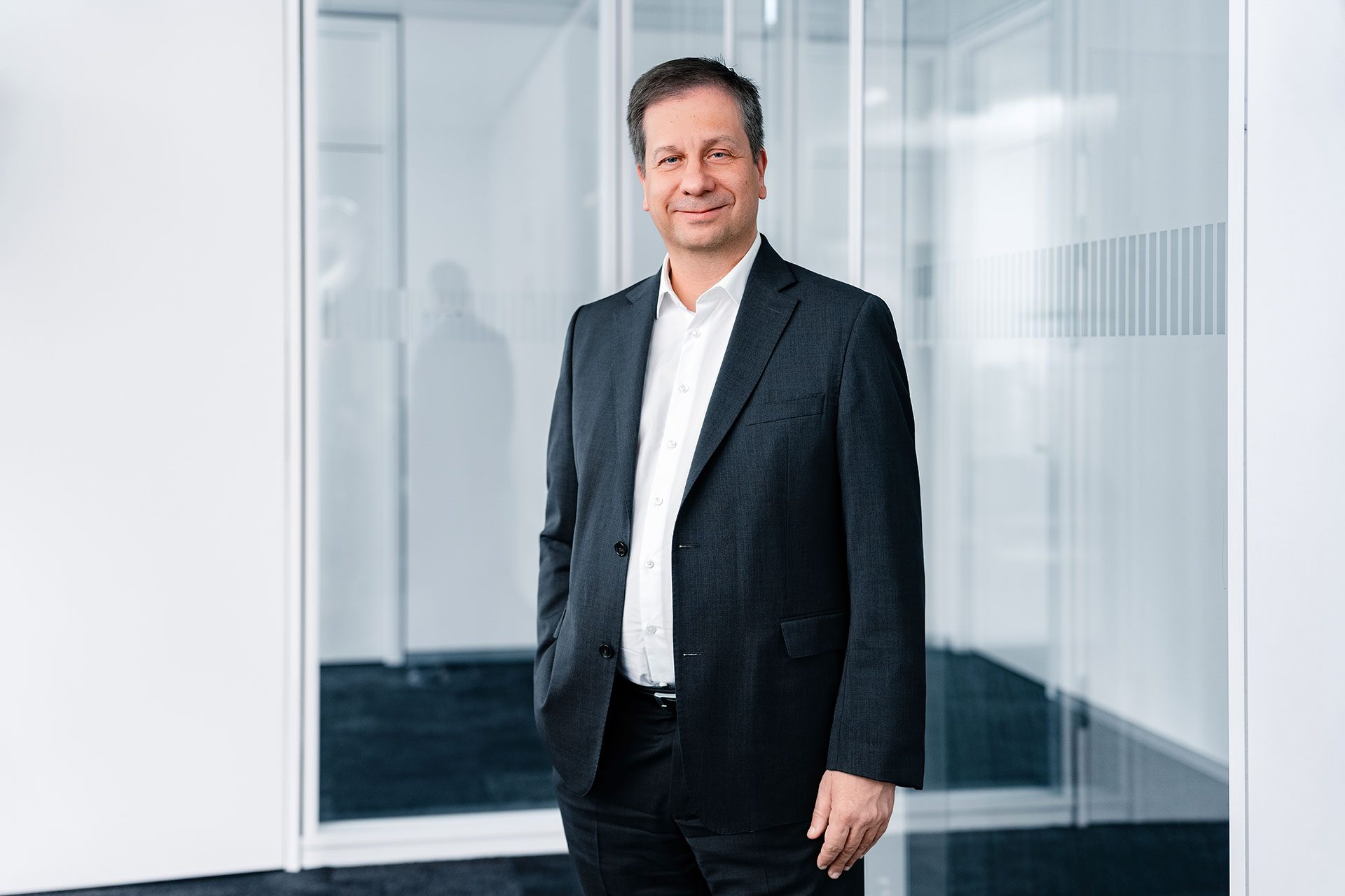 Luka Mucic wearing a dark suit and white shirt standing in a modern, bright office setting with glass walls.