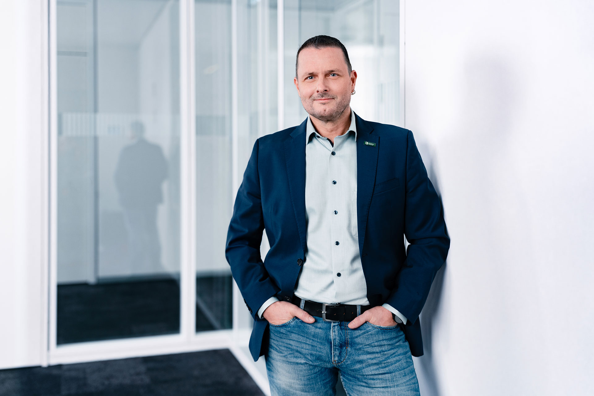 Markus Oleynik wearing a blue blazer, light shirt, and jeans stands casually with hands in pockets near a white wall in an office space with glass partitions in the background.