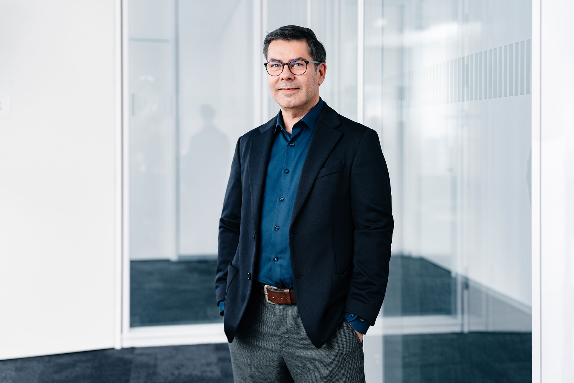 Peter Riedel in business attire, wearing a navy blazer, blue shirt, and gray pants, stands in a modern office space with glass walls.