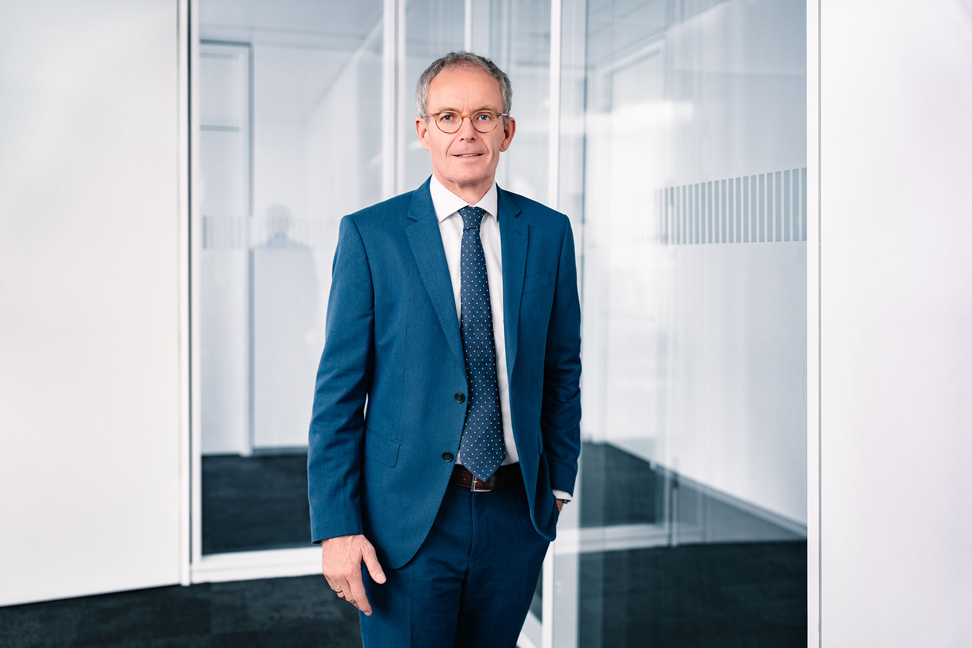 Bernd Scheifele a blue suit and tie stands in a modern, glass-walled office space with black carpet and white walls.