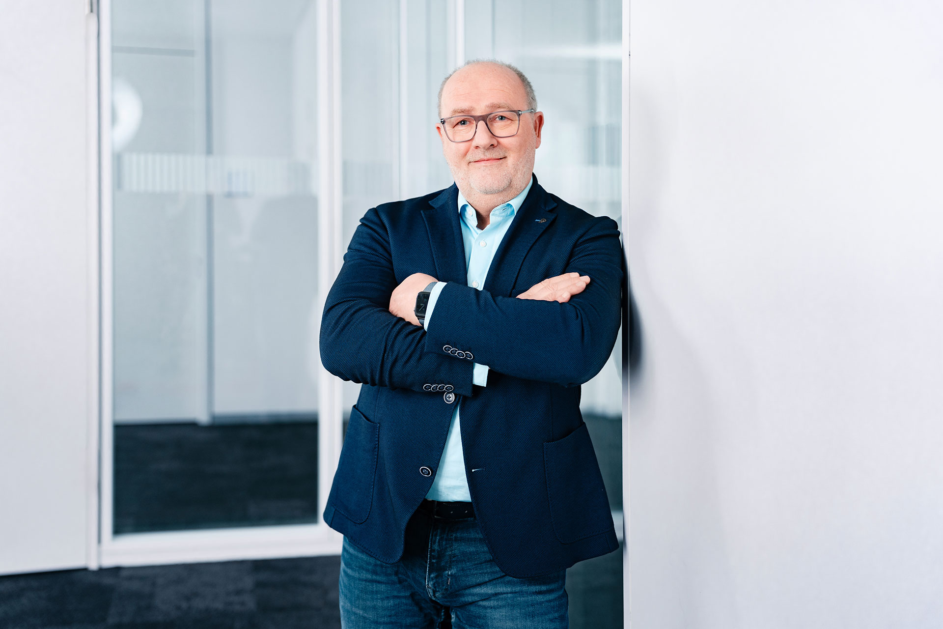 Werner Schraeder stands with arms crossed, leaning against a white wall in a modern office setting with glass partitions.