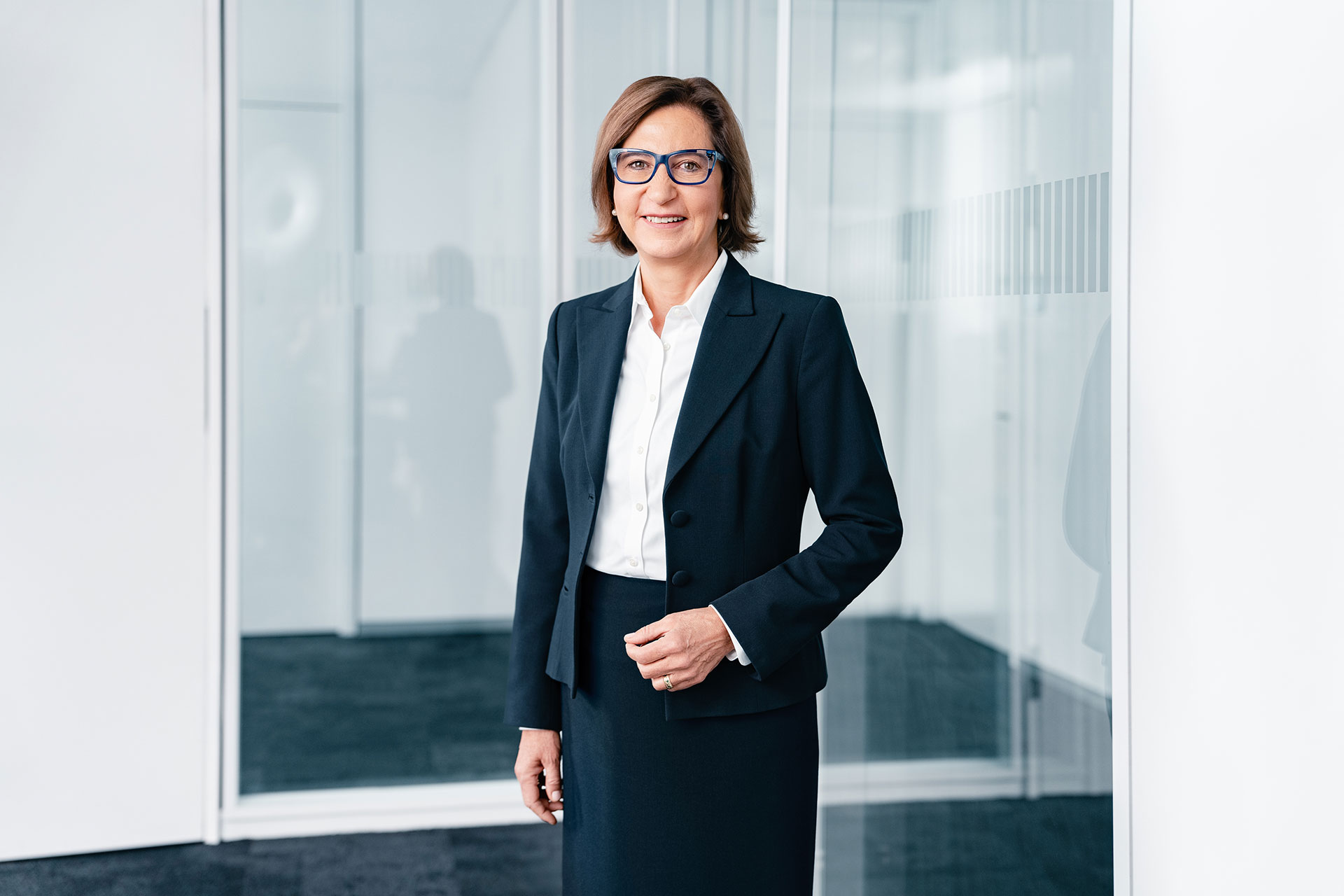 A professional individual wearing a navy business suit and white blouse stands in a modern office setting with glass walls and reflections in the background.