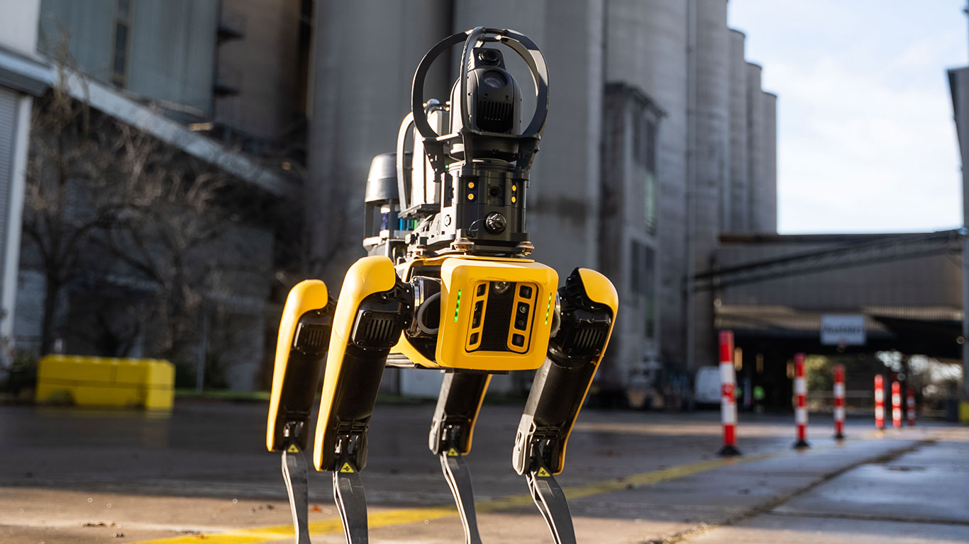 A yellow and black four-legged robotic dog stands on a paved road in an industrial setting.