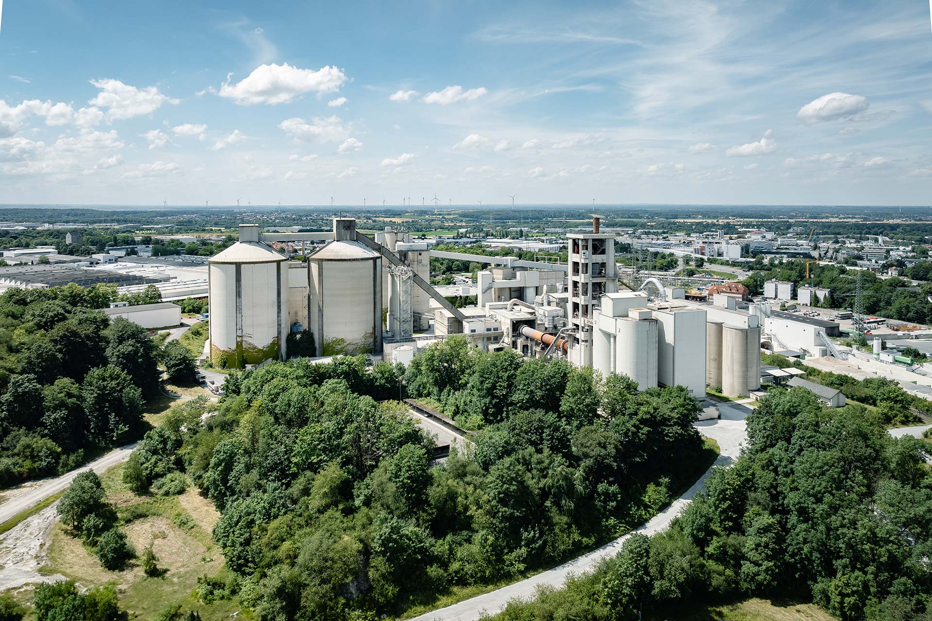 An industrial site with large silos and buildings surrounded by green vegetation, set against a distant urban background.