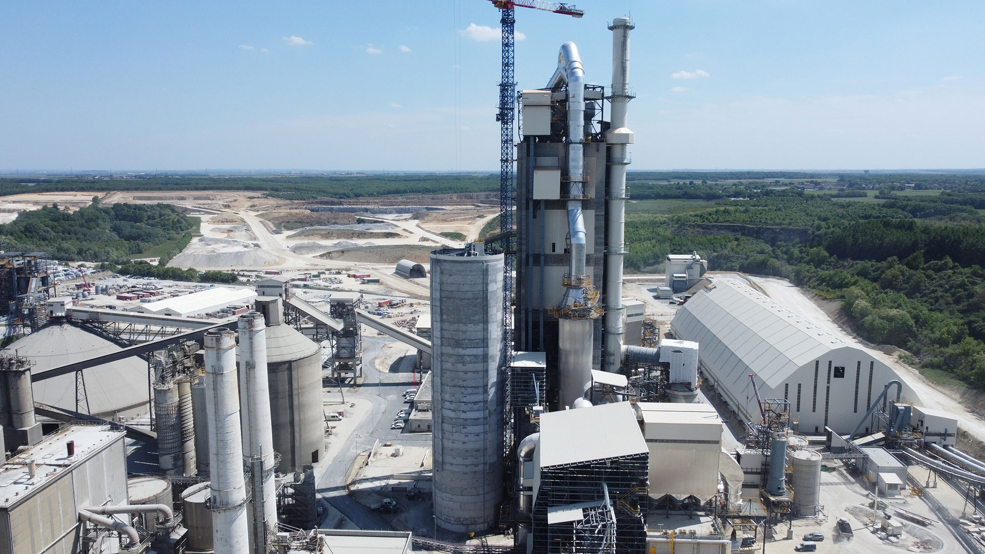 An industrial cement plant with tall silos and surrounding structures, set in a rural area with open land and trees in the background.