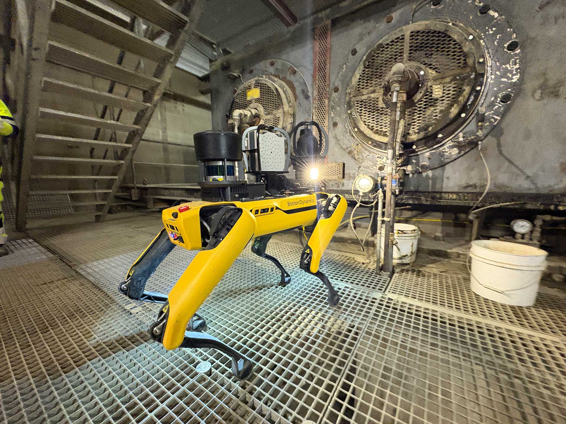 A robotic dog equipped with sensors is inspecting an industrial site with large machinery in the background.