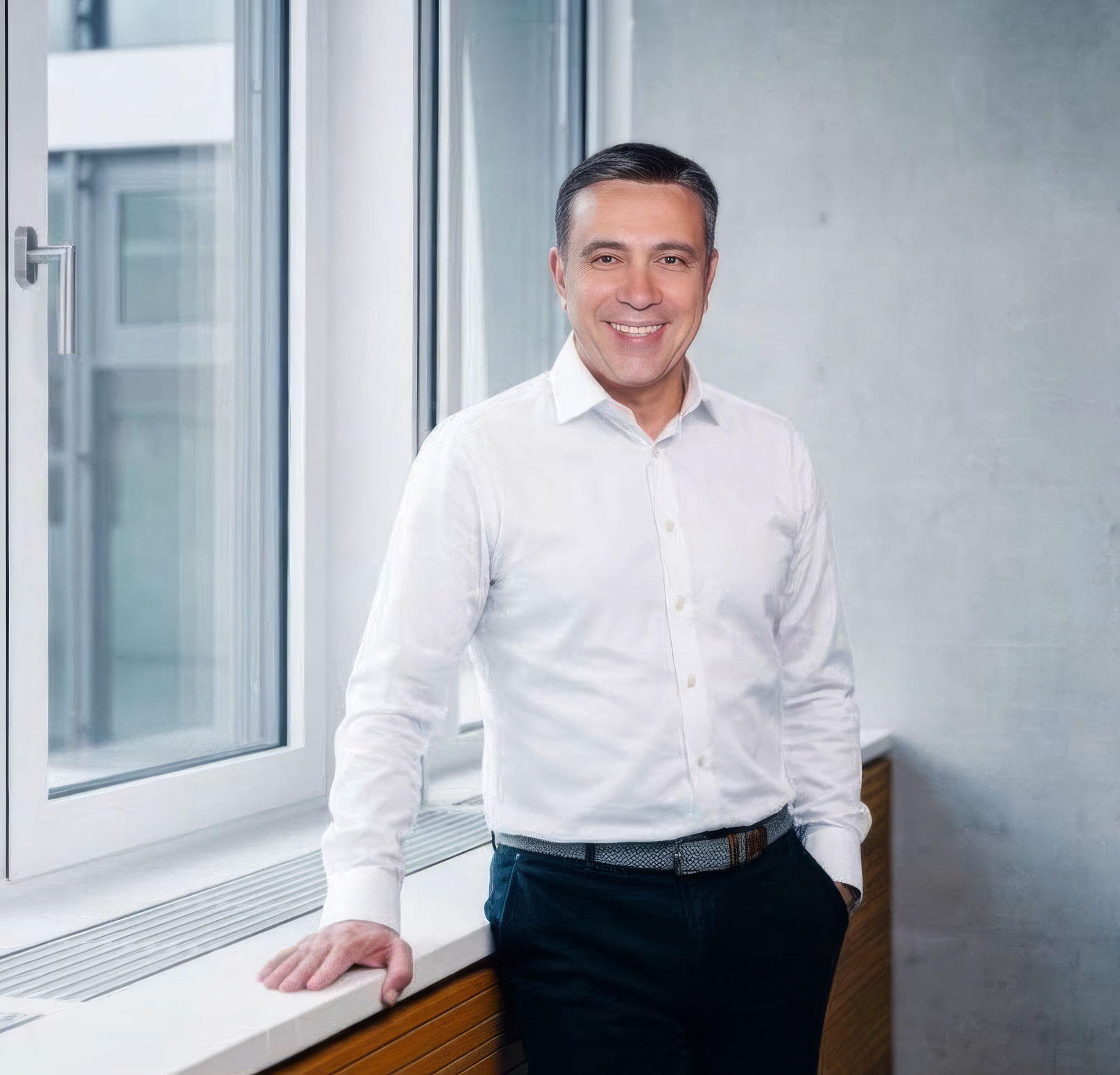 Ozan Kacar wearing a white shirt standing in front of a window, one hand on the windowsill