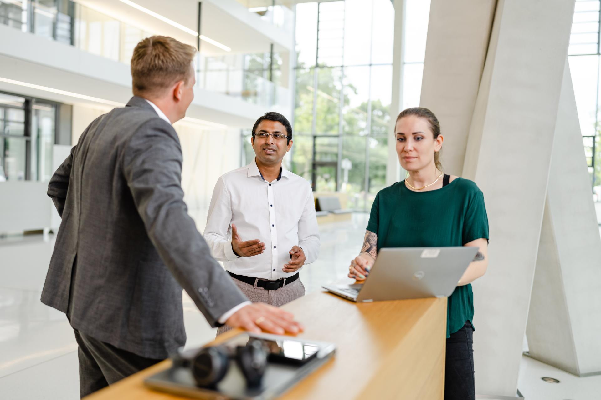 Three colleagues engaged in a discussion in a modern office environment with large windows and natural lighting.