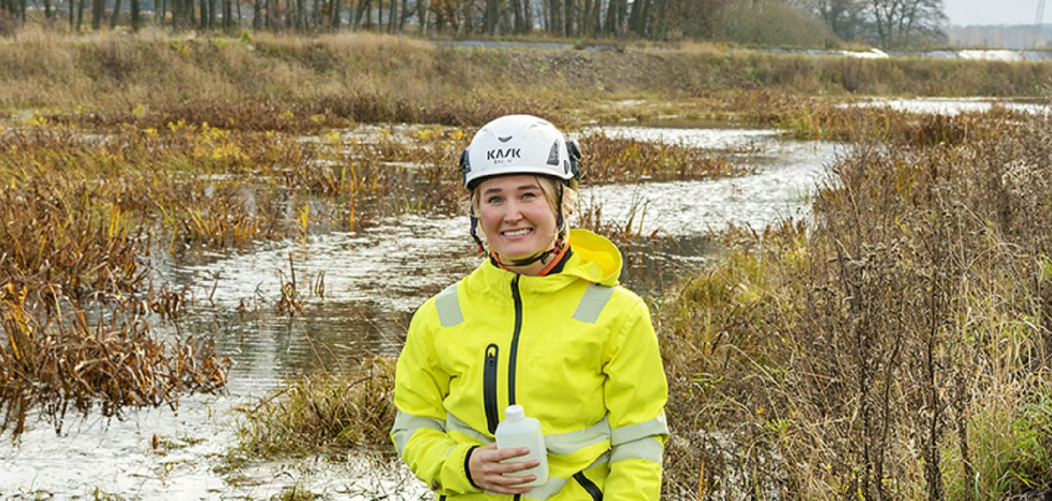 Person in protective clothing stands by a watercourse in a damp, flat, sparsely vegetated landscape