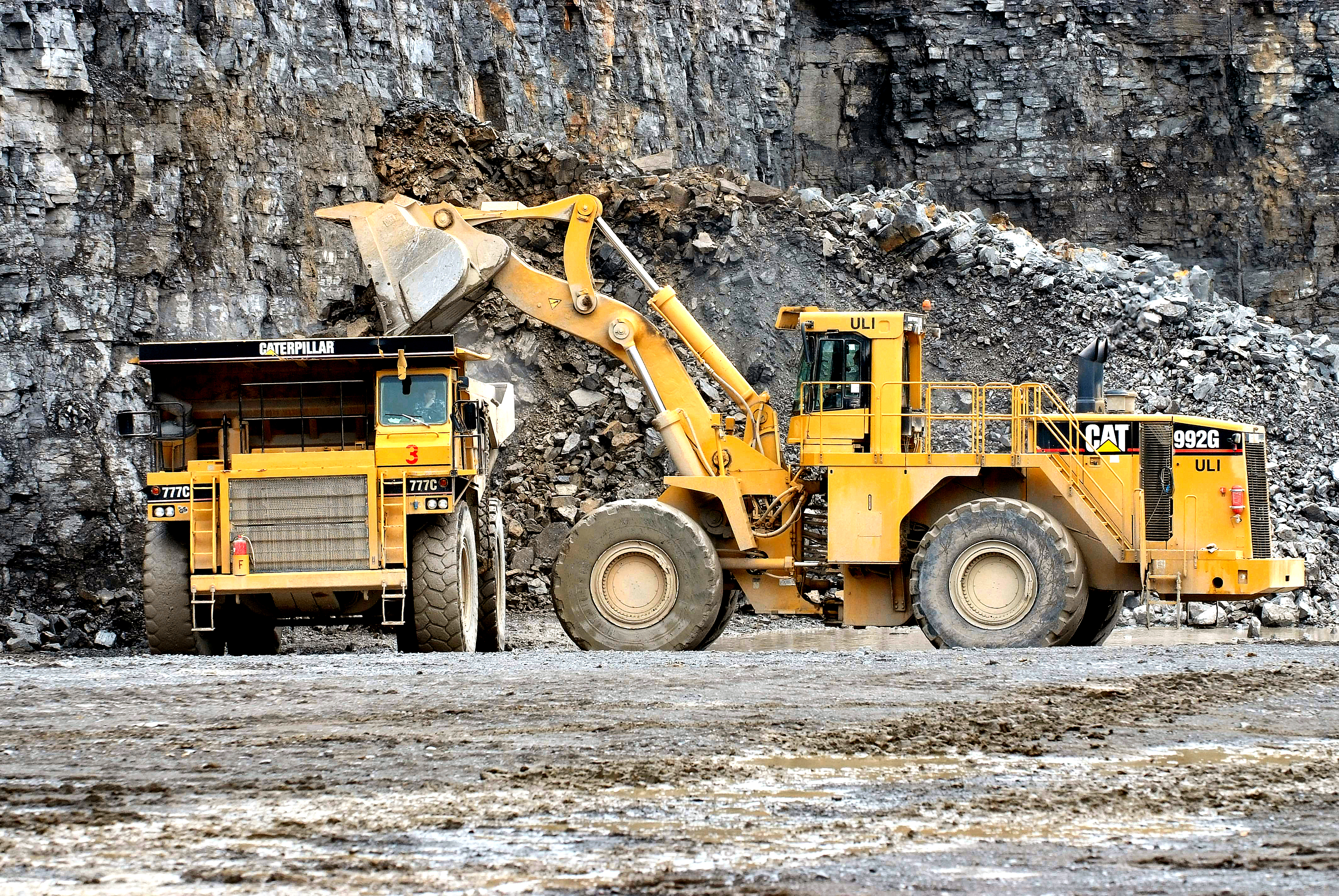 Yellow wheel loader loads dumper truck in front of a stone wall in the quarry