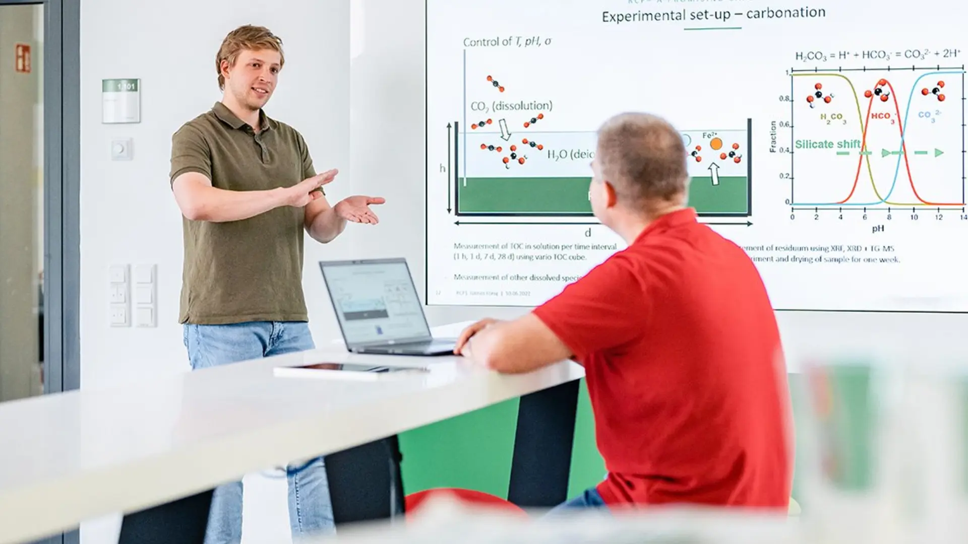 A man standing in front of a white board, explaining something to another person that is sitting on a table