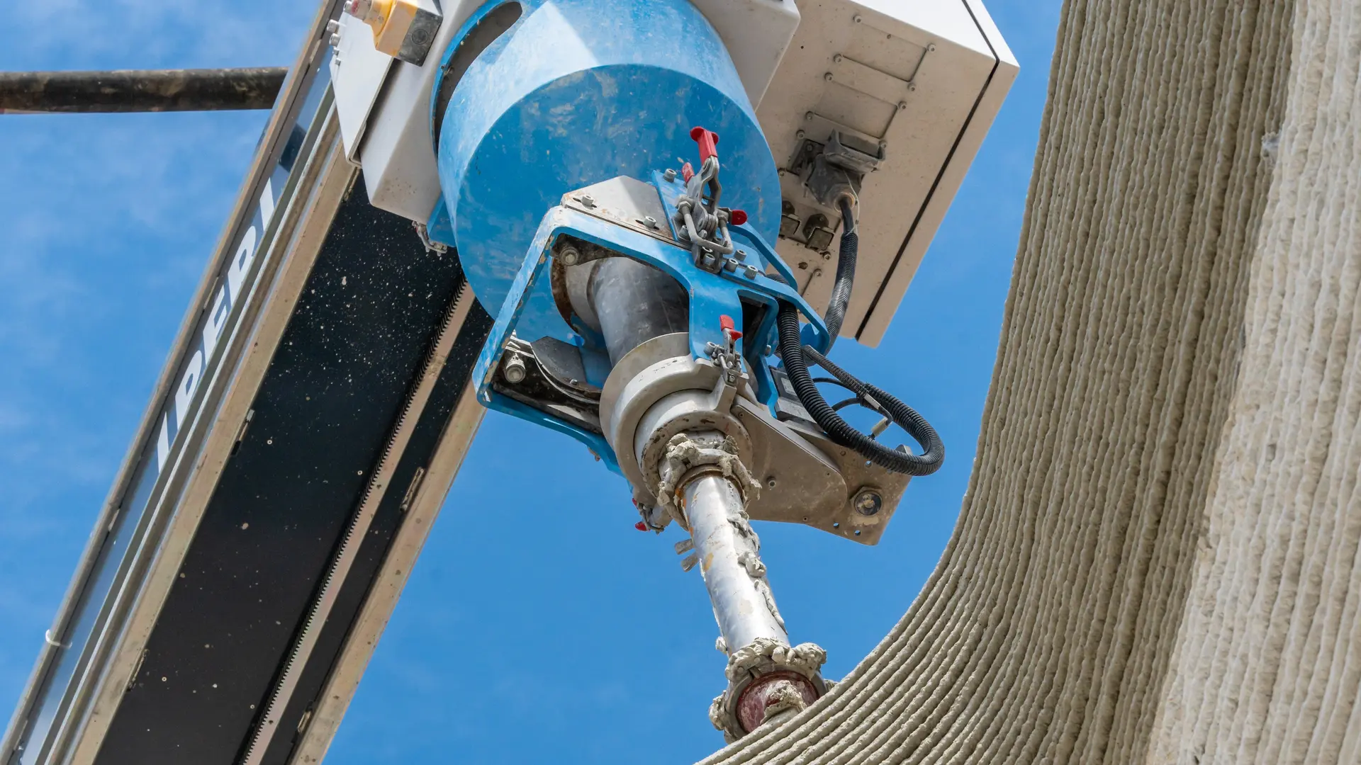 Frog's-eye view: A robot applies a layer of concrete to a 3D-printed wall, blue sky in the background.