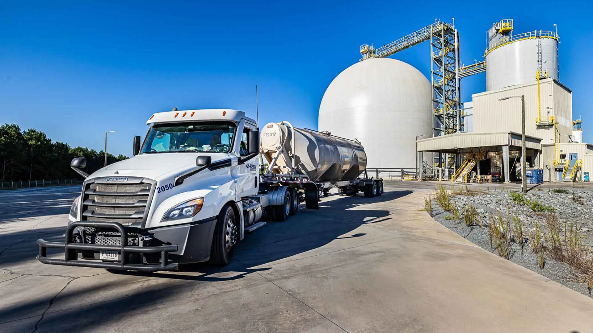 Ein weißer Sattelschlepper parkt auf einem gepflasterten Parkplatz vor einem Zementwerk mit einem großen weißen kuppelförmigen Lagertank, Metallgerüsten und zylindrischen Strukturen unter einem klaren blauen Himmel.