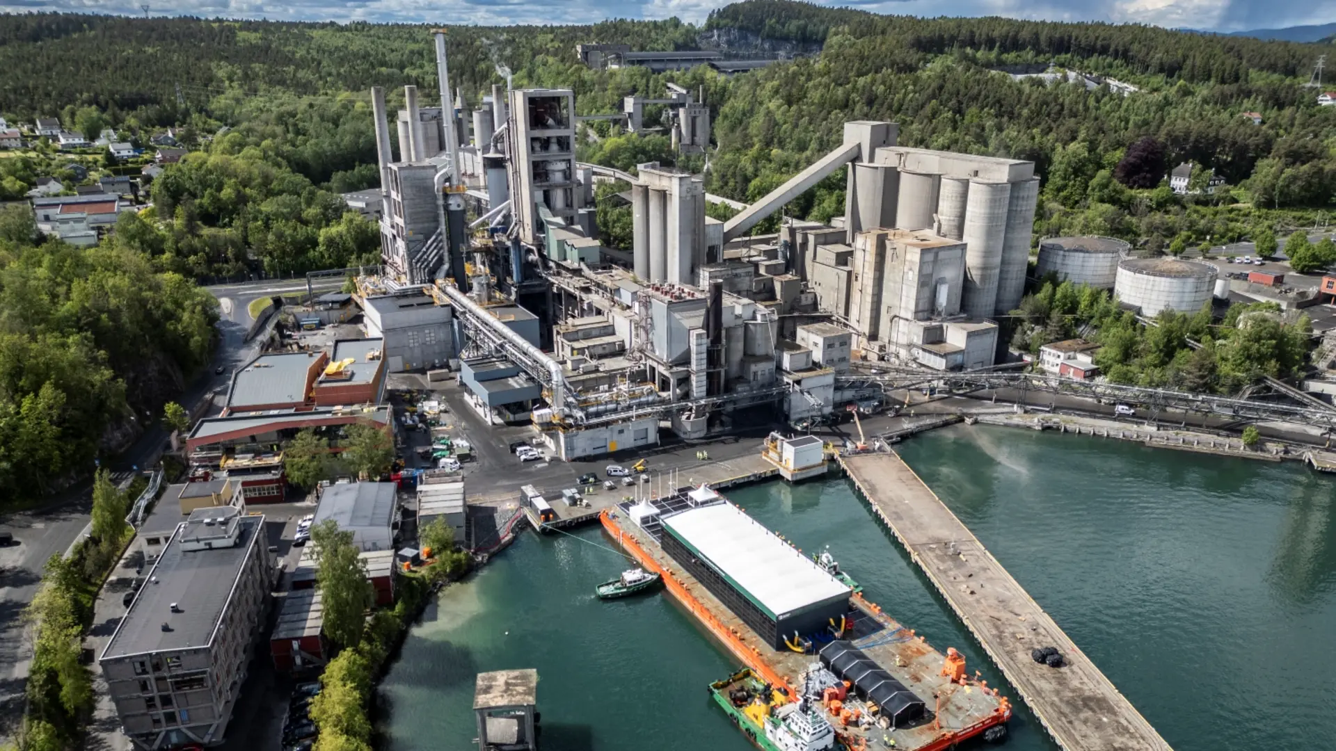 Aerial view of a cement plant with silos, conveyors, and processing units, located near a port with a docked barge for material transport.