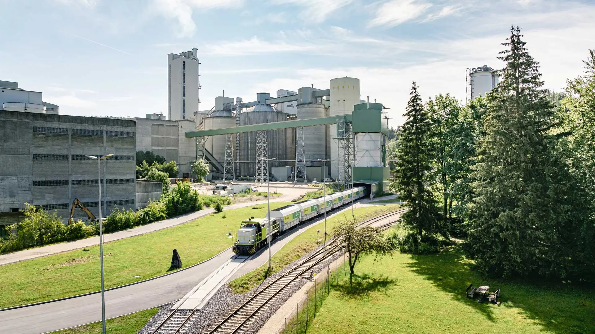 Ein Güterzug fährt auf einer kurvigen Strecke durch eine grüne Landschaft mit Bäumen auf der rechten Seite und einer Zementfabrik mit hohen Silos und Gebäuden im Hintergrund unter einem teilweise bewölkten Himmel.