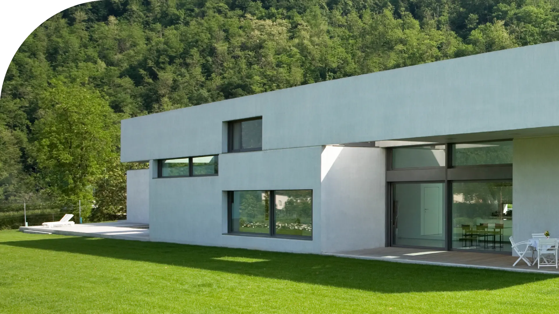 Modernes weißes Flachdachhaus mit klaren, rechteckigen Linien, großen Fenstern und einer Terrasse mit Tisch und Stühlen. Das Haus ist von einer grünen Wiese umgeben, im Hintergrund sind Wald und Hügel sichtbar.