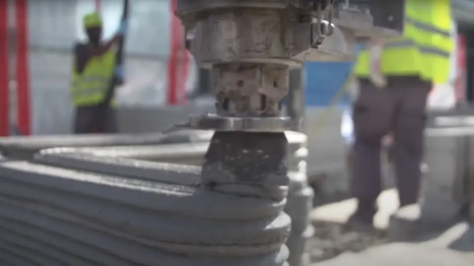 Detailansicht eines 3D-Betondruckers, der gerade eine weitere Schicht Beton auf eine Mauer aufträgt. Im Hintergrund steht eine Person in Schutzkleidung.