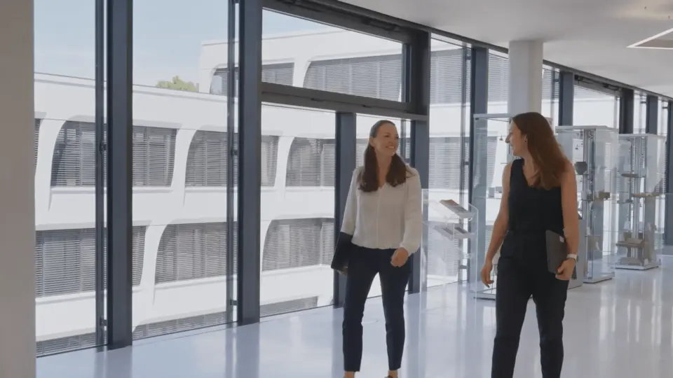 Two women walking and conversing in a bright, modern office hallway with large floor-to-ceiling windows on the left, revealing an exterior view of a white building. One woman holds a laptop.