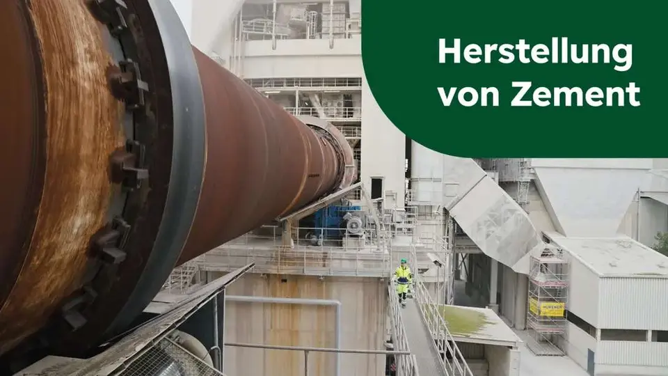 Blick auf ein Zementwerk, links ein Drehofen, unterhalb an einem Geländer ein Arbeiter in Schutzkleidung mit grünem Helm; oben rechts steht auf grünem Grund "Herstellung von Zement"