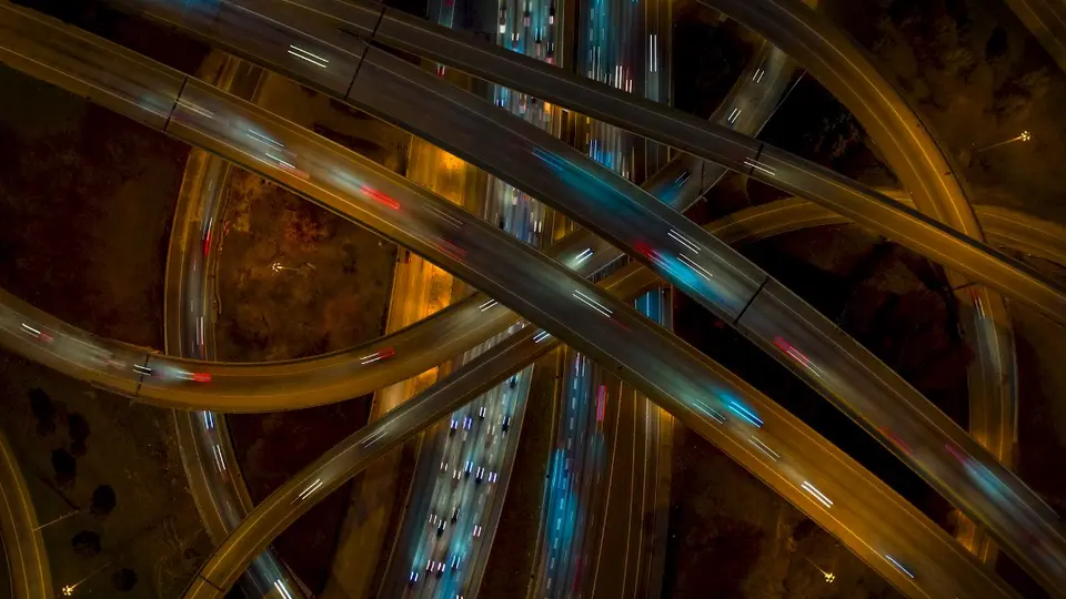 Aerial view of a complex multi-level highway interchange at night, illuminated by streetlights. Traffic flows along various curved and intersecting roadways, with red and white car lights visible in opposite directions.