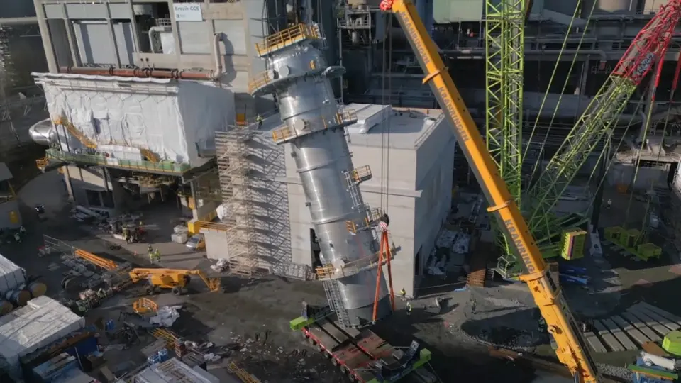 A large industrial construction site with a cylindrical steel structure being lifted into place by a yellow crane on the right. Surrounding the structure are workers in safety gear, scaffolding, concrete buildings, and various construction equipment. The area is cluttered with tools, materials, and machinery.
